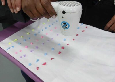 Hands using a small hair dryer to move colorful paint dots on a white surface.