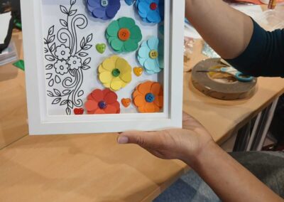 Handmade paper flower art in a white shadow box frame with colorful buttons and drawn floral accents.