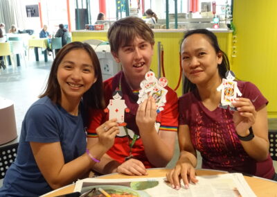 Three smiling people holding paper dolls at a table in a cafe.