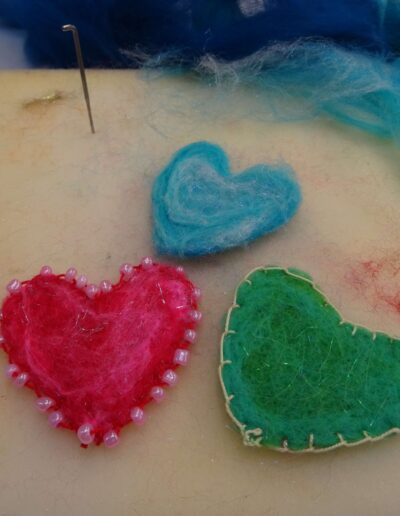 Needle felted hearts in blue, pink with beads, and green on a foam pad with felting needle and blue roving.