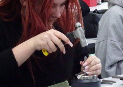 Young woman hammering metal for jewelry making, wearing red-framed glasses.