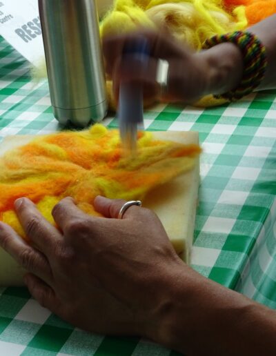 Needle felting wool: Hands crafting a sun-like design with yellow and orange wool on a foam pad.