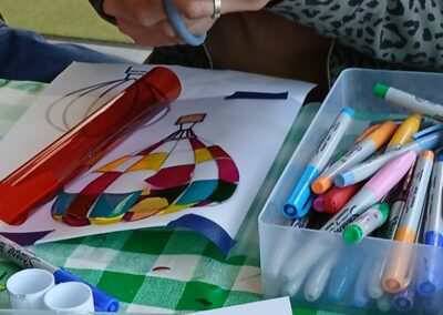 Craft project: Hands cutting red plastic near a colorful hot air balloon drawing and markers.